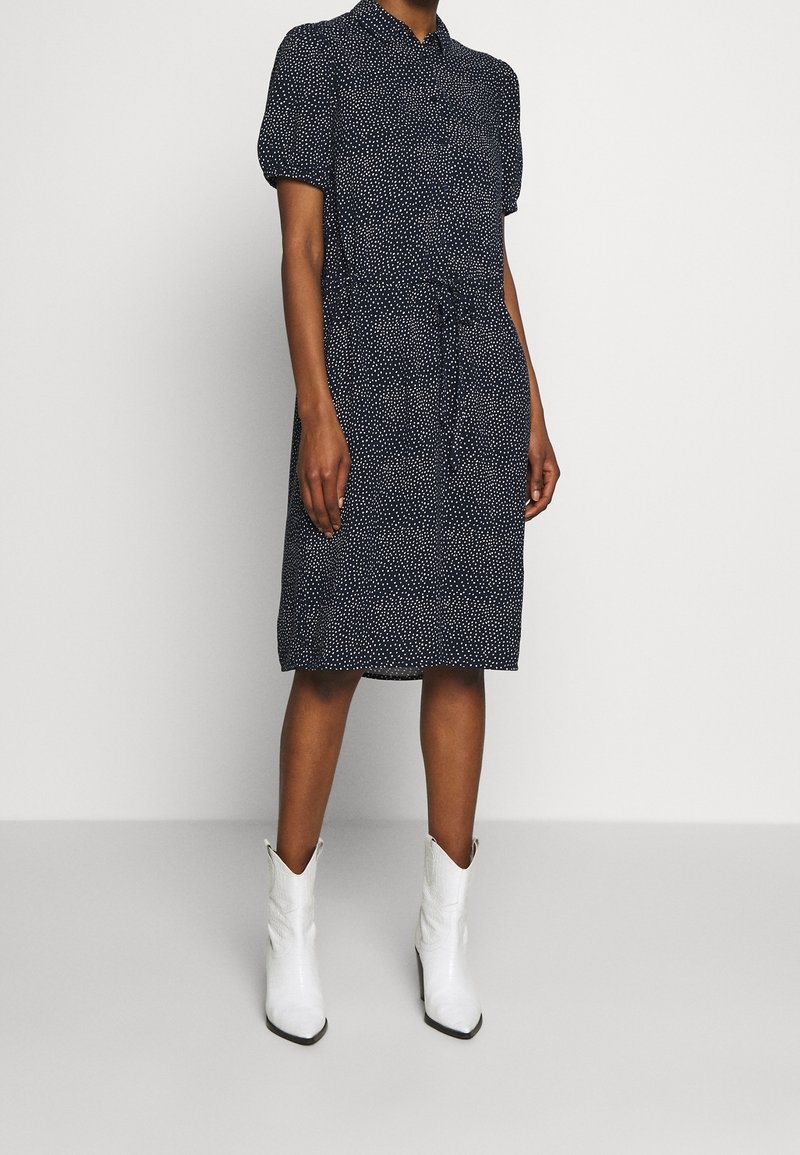 Femme portant une robe bleu marine longueur genou avec des pois blancs et des manches bouffantes, associée à des bottines blanches à bout pointu.
