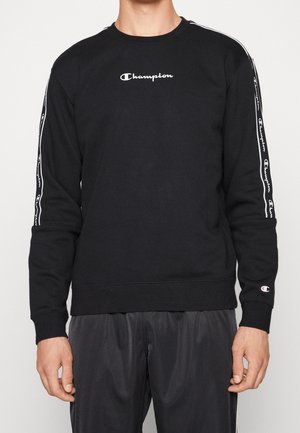 Personne portant un sweat-shirt Champion noir avec un logo blanc sur la poitrine, un ruban à logo sur les manches et un patch logo sur le poignet gauche.