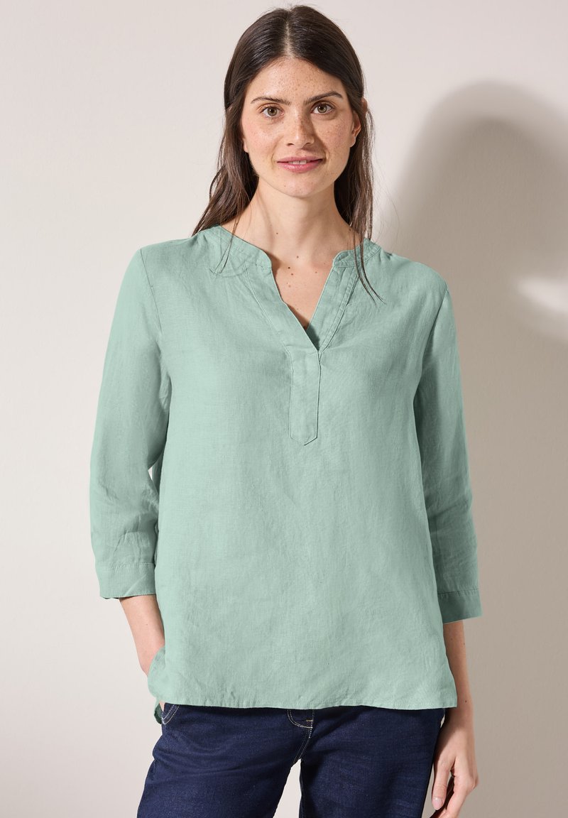 Blusa in lino verde chiaro con scollatura a V, maniche a tre quarti e vestibilità rilassata. Caratteristiche notevoli includono un tessuto leggermente texturizzato e un orlo curvo.