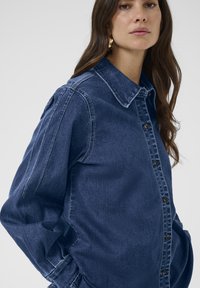 Sötétkék farmering klasszikus gallérral, elöl gombos záródással és enyhén puffos ujjakkal. Kontrasztos varrással és laza szabással rendelkezik.