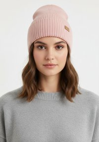 Lichtroze gebreide muts met geribbelde textuur en gestikte details, met een klein bruin leren label; informeel gedragen over bruin haar.