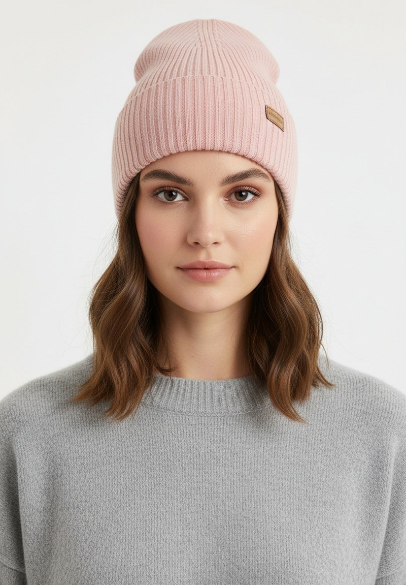 Lichtroze gebreide muts met geribbelde textuur en gestikte details, met een klein bruin leren label; informeel gedragen over bruin haar.