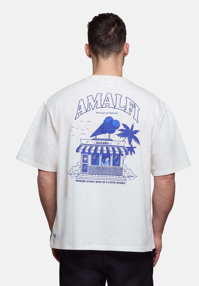Homme portant un T-shirt blanc avec une illustration bleue de la boutique de gelato Amalfi, un palmier, et le texte « Où chaque bouchée est une histoire d'amour. »
