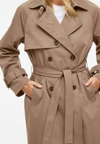 Trench-coat beige en tissu lisse, doté d'une coupe croisée, d'un col large, d'une ceinture et de boutons comme détails.