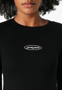 Svart ribbet langermet topp med en oval "guess jeans" logo i hvit broderi på brystet. Jevn tekstur med en tettsittende design.