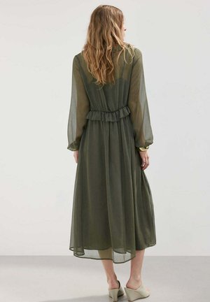 Vrouw met lang golvend haar draagt een doorschijnende olijfgroene midi-jurk met lange mouwen en ruches in de taille, beige sleehakken en een gouden armband.