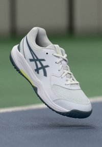 White sneaker with a textured upper, grey accents, and a greenish-yellow sole. Features a lace-up closure and a rounded toe design.