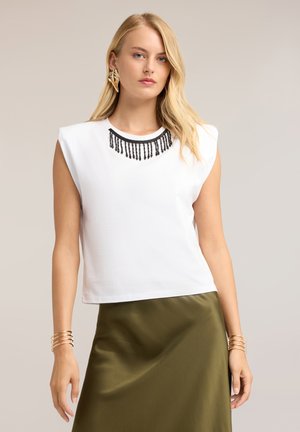 Blonde vrouw die een mouwloos wit topje draagt met zwarte kralen aan de halslijn en een olijfgroene rok, staand voor een neutrale achtergrond.