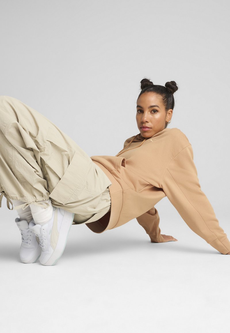 Felpa oversize beige abbinata a pantaloni jogger beige chiaro a gamba affusolata e sneakers bianche, su uno sfondo grigio uniforme.