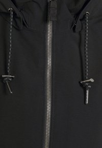 Svart jacka med en silverdragkedja, justerbara snören och ett slätt, texturerat tyg. Funktionella detaljer förbättrar designen.