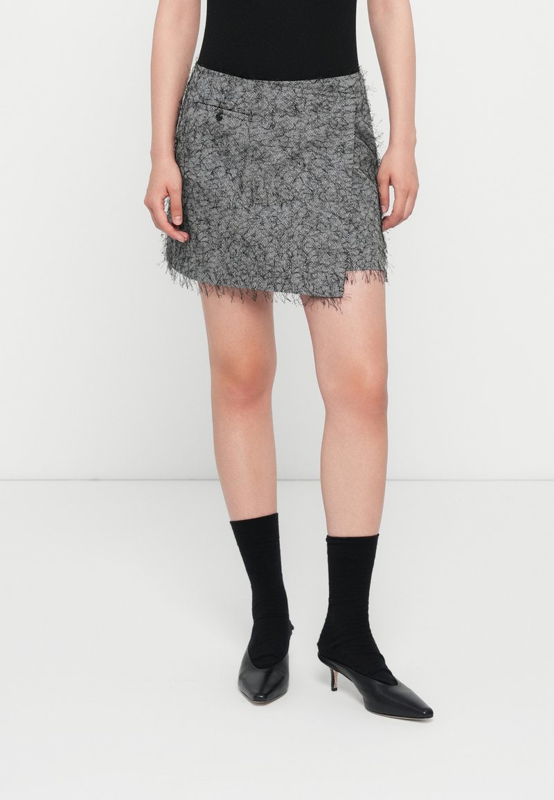 Grauer gewebter Minirock mit franstem Saum, ausgestattet mit einer Seitentasche und strukturiertem Muster. Kombiniert mit schwarzen Socken und spitzen Absatzschuhen.