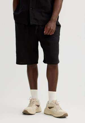 Hombre vestido con pantalones cortos negros, calcetines blancos y zapatillas beige, de pie frente a un fondo liso.