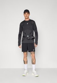 Black long-sleeve athletic shirt with textured design, paired with black shorts. A water bottle is secured in a belt. White sneakers with green accents.