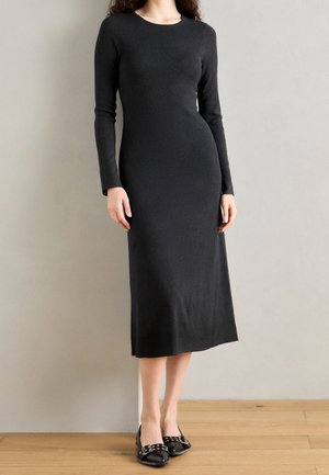 Femme portant une robe midi noire ajustée à manches longues et des ballerines noires avec des détails à boucle argentée, debout contre un mur clair uni.