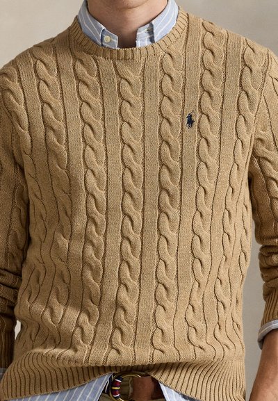 Man draagt een lichtbruine kabelgebreide trui over een blauw gestreept overhemd met knopen, met een bruine riem zichtbaar bij de taille.