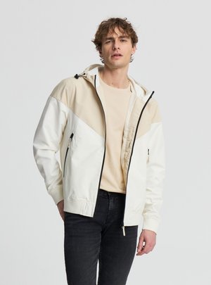 Giovane uomo che indossa una giacca con cappuccio beige e bianca con zip sopra una camicia beige chiaro e jeans neri, in piedi davanti a uno sfondo semplice.