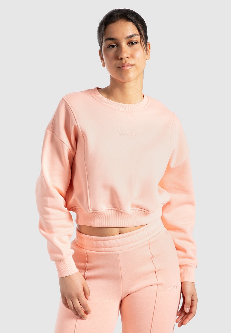 Jasnoróżowy skrócony sweatshirt z okrągłym dekoltem, ściągaczami przy mankietach i u dołu. Połączony z pasującymi zwężającymi się spodniami, oba wykonane z miękkiego materiału.