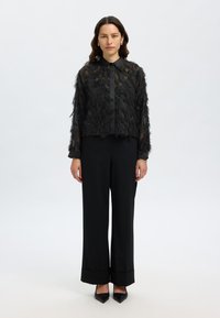 Black sheer top with textured, layered fringe, collared, long sleeves; paired with tailored black trousers and pointed-toe heels.