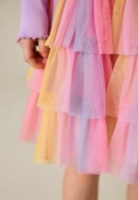 Main d'enfant à côté d'une jupe en tulle superposée aux teintes pastel rose, violet et jaune, portant un haut à manches longues lavande avec des poignets à volants.