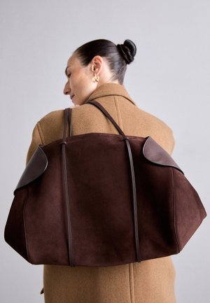 Bolso de ante marrón con acentos de cuero oscuro, que presenta una forma estructurada y dos asas, sostenido sobre el hombro contra un abrigo beige.