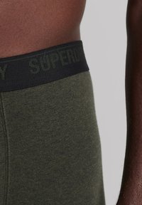 Boxers en coton vert olive avec une ceinture élastique noire arborant le logo "SUPERDRY" en vert foncé. Texture lisse et coupe ajustée.