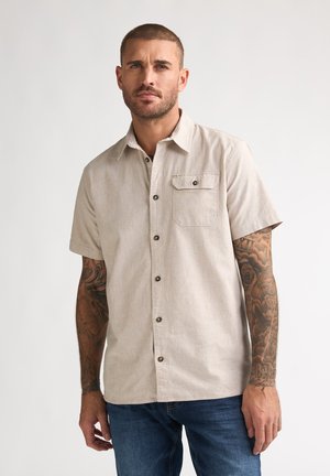 Mann mit kurzen Haaren und Tätowierungen an beiden Armen, der ein beige kurzärmeliges Hemd mit Knöpfen und blaue Jeans vor einfachem Hintergrund trägt.
