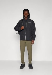 Schwarze wasserdichte Jacke mit Kapuze, Reißverschluss und elastischen Bündchen; kombiniert mit olivfarbenen Hosen und schwarzen Wanderschuhen. Robuster Stoff und Design.