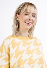 Sárga és fehér mintás pulóver, amelyet houndstooth minta díszít, kötött textúrával és kerek nyakléccel.
