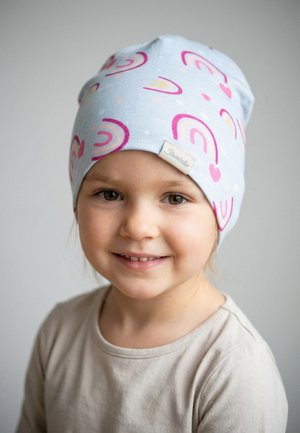 Young child with light beige shirt wearing a light blue beanie decorated with pink and purple rainbows and hearts, smiling softly.