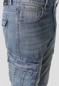 Pantalon cargo en denim bleu clair avec plusieurs poches, coutures apparentes et marquage « KAS DENIM KOROSHI » sur la zone de la poche avant.