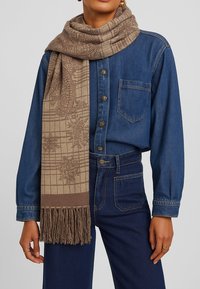 Sciarpa beige con un motivo a griglia testurizzato e bordi frangiati, posata su una camicia di denim con dettagli sui bottoni e una vestibilità classica.