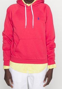 Felpa rossa di cotone con cordini bianchi, tasca a marsupio e logo ricamato blu. Indossata sopra una maglietta gialla pallida.