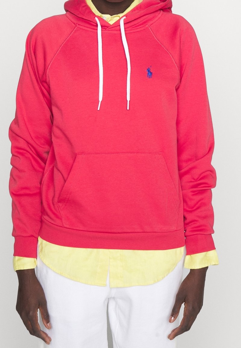 Felpa rossa di cotone con cordini bianchi, tasca a marsupio e logo ricamato blu. Indossata sopra una maglietta gialla pallida.