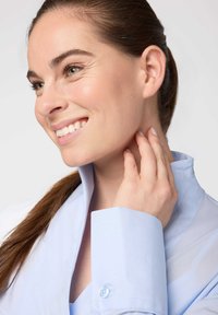 Camicia azzurra chiara con colletto alto, polsini abbottonati e texture liscia. Capelli raccolti, che mettono in evidenza la pelle chiara e il trucco naturale.