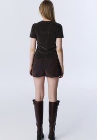 Vrouw die met haar rug naar de camera staat, gekleed in een zwart short-sleeve top, donkerbruine shorts en knielange donkerbruine laarzen, tegen een effen achtergrond.