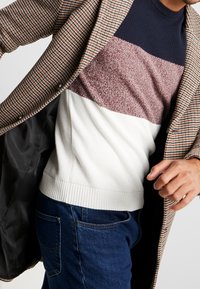 Hombre vestido con un suéter de rayas multicolores, jeans azules y un abrigo de patrón de cuadros marrón, sosteniendo el abrigo abierto con una mano.