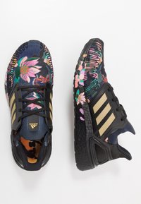 Zapatillas deportivas negras con un patrón floral, con parte superior de punto, suela texturizada negra y tres rayas doradas. Cuenta con un sistema de cordones para mayor soporte.