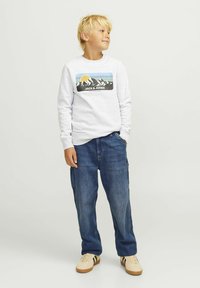 Svijetlosiva dukserica s grafikom planine i natpisom "JACK & JONES", uparena s plavim trapericama sa džepovima i bež tenisicama sa prugama.