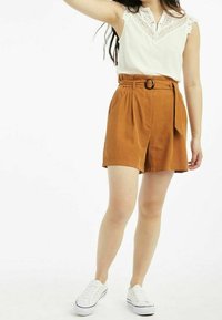 Bílá bezramínková blůza s krajkovým lemem, zvolená k šortkám v barvě rezavé s vysokým pasem, které mají skladby a pásek, nošeno s bílými teniskami.