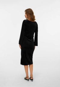 Vestido midi de terciopelo negro con mangas largas, espalda descubierta y silueta ajustada, combinado con sandalias de tacón en blanco y negro.