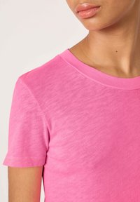 Primo piano di una persona che indossa una T-shirt rosa acceso a maniche corte con scollo rotondo, mostrando collo, spalla e parte superiore del braccio.
