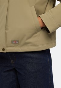 Giacca beige con una texture liscia, caratterizzata da una patch con logo visibile, tasche laterali e cuciture verdi a contrasto, abbinata a pantaloni di jeans scuri.