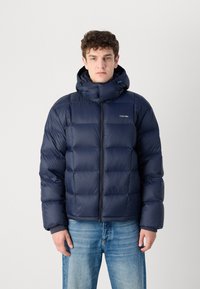 Navy puffer jacket with a high collar, zip front, and elastic cuffs. Smooth metallic fabric, rectangular quilted design, branded logo on chest.