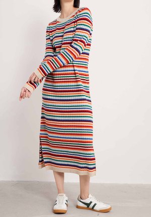 Personne portant une robe midi à manches longues rayée multicolore et des baskets blanches avec des bandes vertes, debout sur un sol en béton.