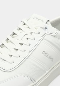 Valge nahast Calvin Klein tennisejalats, millel on valged nöörid, tekstureeritud külgpaneel ja reljeefne brändi logo küljel ja keelel.