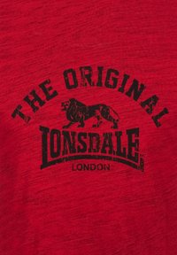 Piros póló, puha anyagból, merész fekete grafikával egy oroszlánról, valamint a „THE ORIGINAL LONSDALE LONDON” felirattal, ívelt elrendezésben.