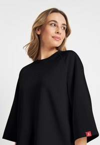 Zwarte oversized top met een ronde hals en driekwart mouwen. Getextureerde stof met een rode labeldetail op de mouw.