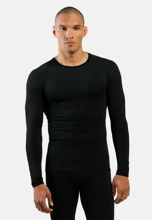 Athletischer Mann mit kurzen Haaren, der ein eng anliegendes schwarzes Langarmshirt und schwarze Hose trägt, vor einem schlichten hellen Hintergrund stehend.