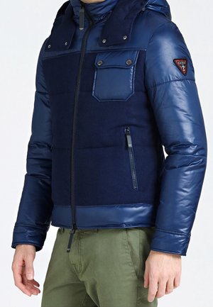 Marineblauwe pufferjack met een mix van glanzende en matte texturen. Voorzien van een rits aan de voorkant, drukknoopsluiting, borstzak en zijzak met rits.