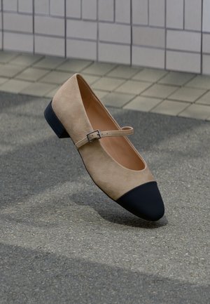 Beige ruskind Mary Jane-sko med sort afrundet snude og lille spænde, vist på en belagt udendørs overflade.